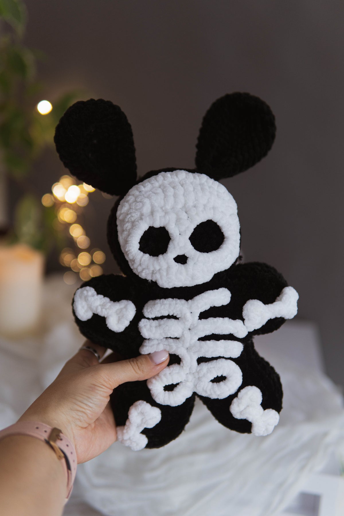 A hand holds a black and white crocheted plush toy with bunny ears and a skeleton design, set against a softly blurred indoor background with warm lights.