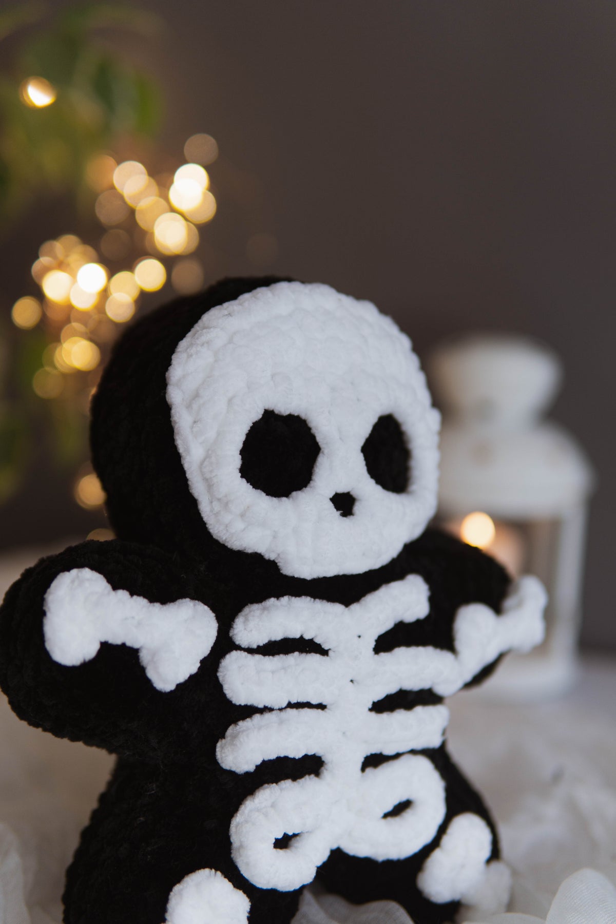 A black and white crocheted plush toy shaped like a skeleton, standing against a softly blurred background with warm bokeh lights and a white lantern.