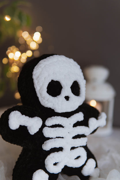 A black and white crocheted plush toy shaped like a skeleton, standing against a softly blurred background with warm bokeh lights and a white lantern.
