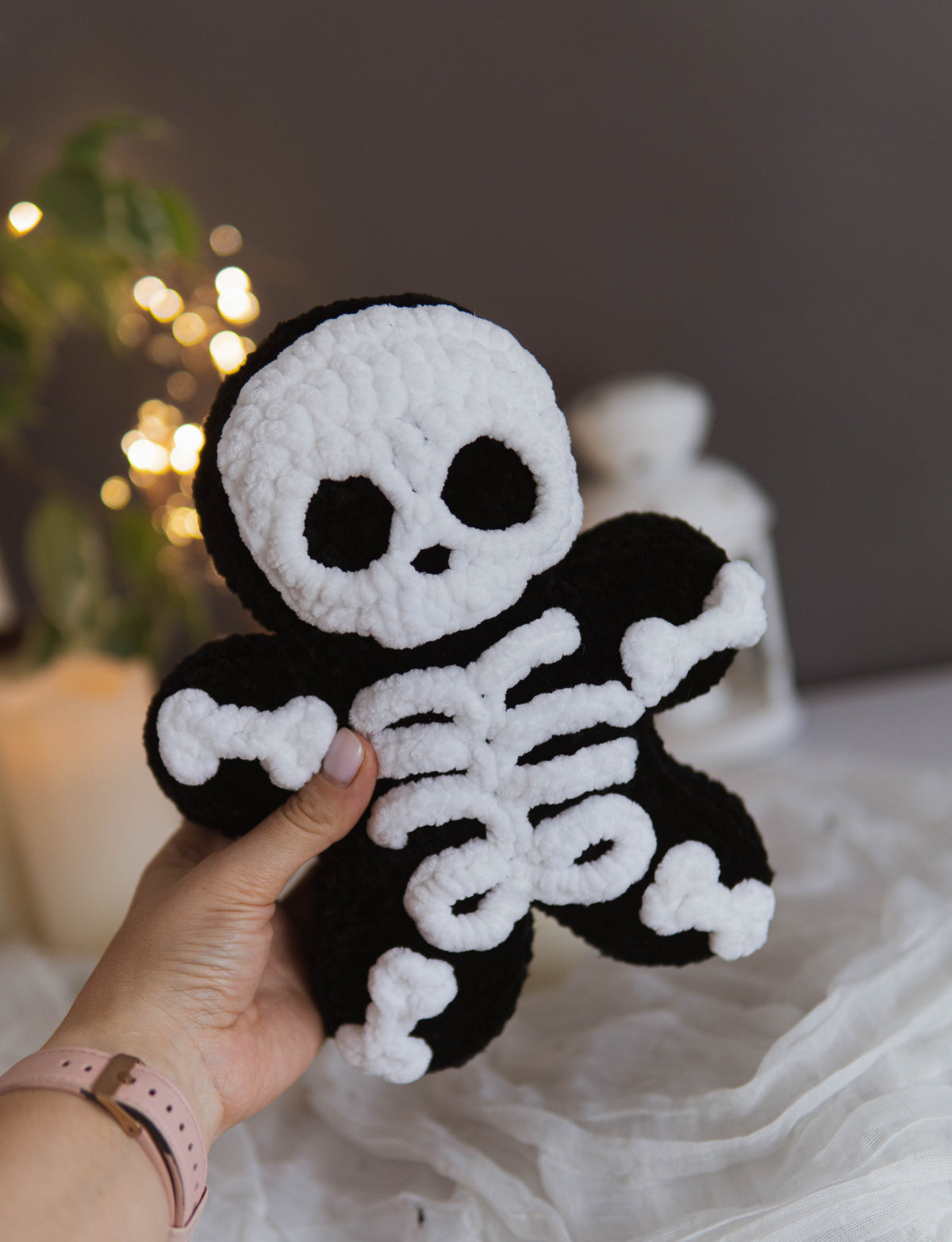 A hand holds a black and white crocheted skeleton plush toy against a softly blurred indoor background with warm lights and white fabric.