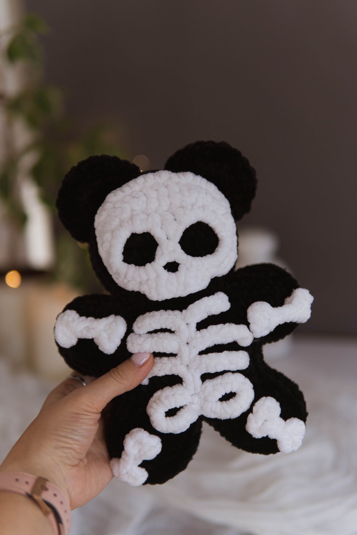 A hand holds a crocheted plush toy shaped like a panda with a white skeleton print on a black background, set against a softly blurred indoor backdrop.
