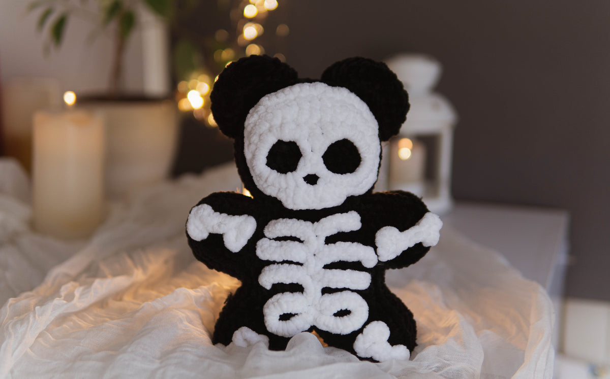 A black and white crocheted skeleton bear plush toy sitting on a white fabric surface with a softly blurred background featuring candles and warm lights.