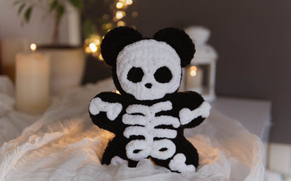 A black and white crocheted skeleton bear plush toy sitting on a white fabric surface with a softly blurred background featuring candles and warm lights.
