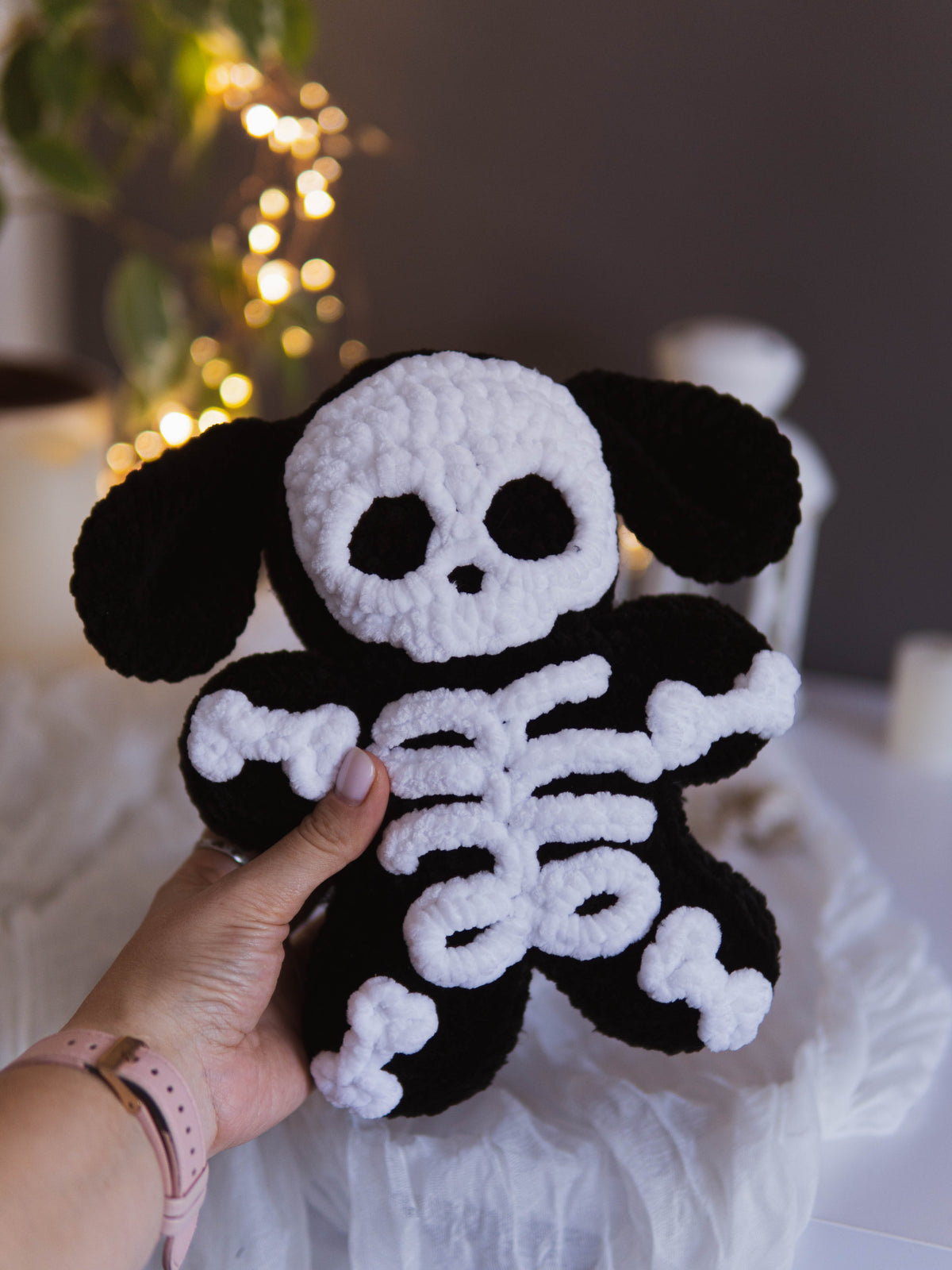 A handmade crocheted plush toy shaped like a skeleton dog, held by a hand. The toy features a white skull and white rib cage over a black body and floppy black ears, set against a softly blurred background with warm bokeh lights.