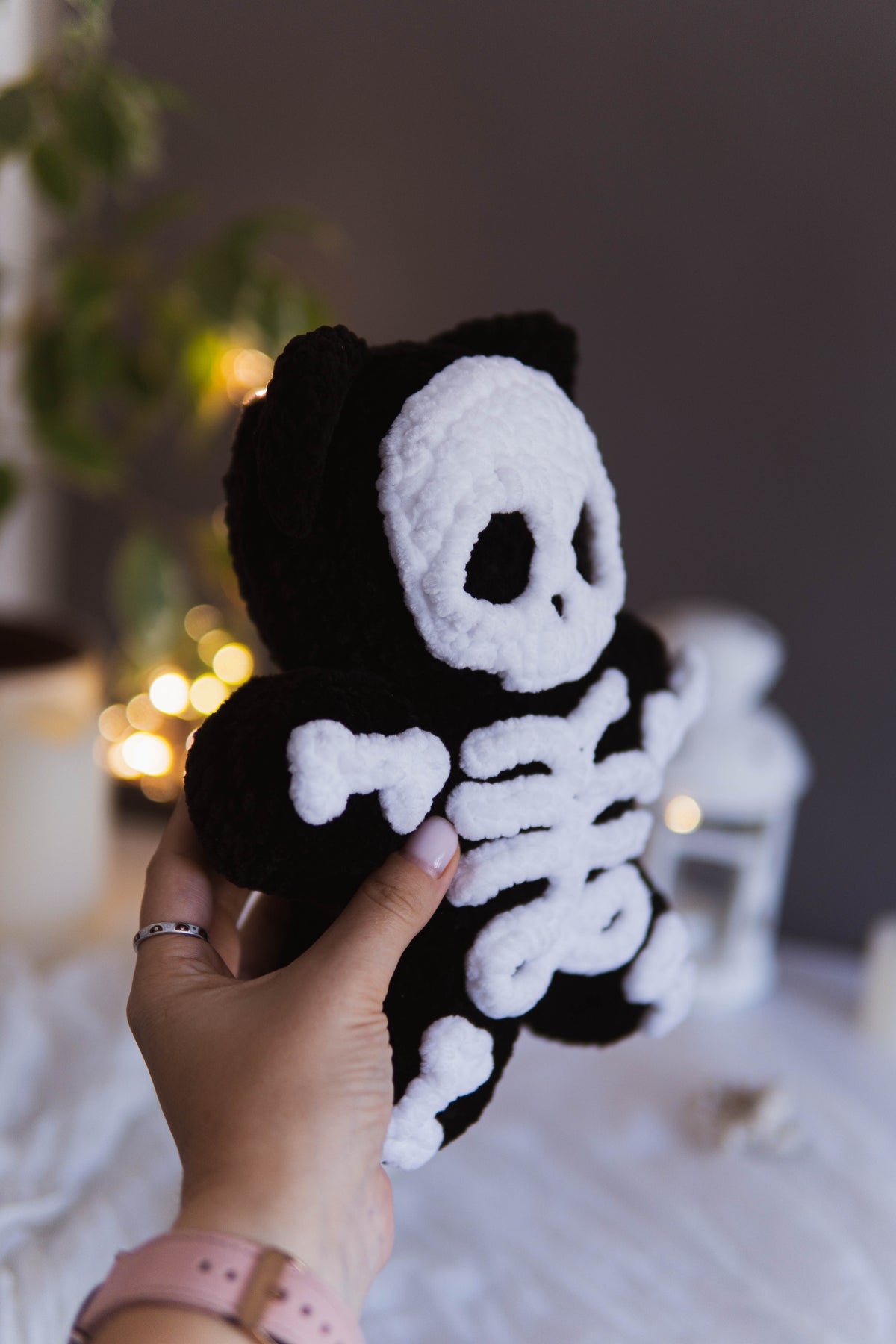 A hand holds a black and white plush skeleton toy against a softly blurred indoor background with warm lights and white decor.
