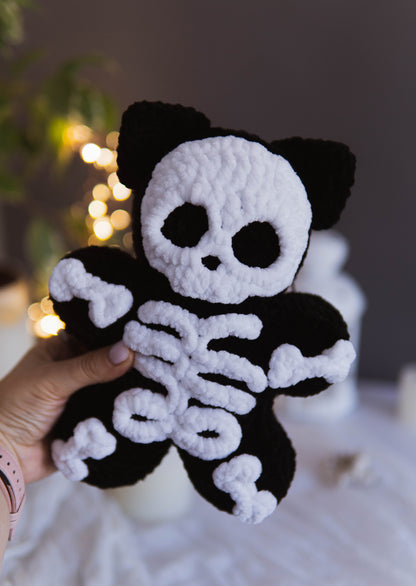 A hand holds a black and white crocheted plush toy shaped like a skeleton cat, with a skull face, cat ears, and white bones on a soft, blurred indoor background.