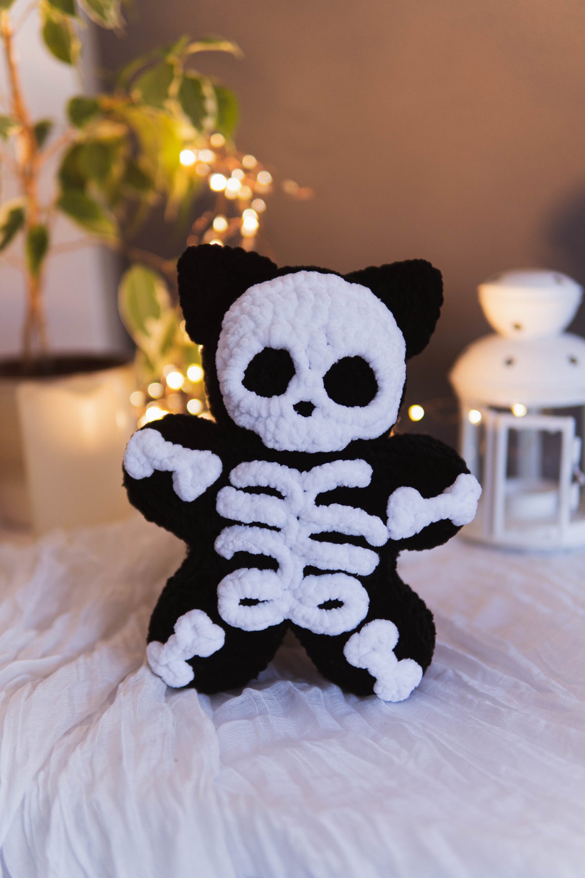 A black and white crocheted plush skeleton cat toy standing on a white fabric surface with a warm, softly blurred indoor background featuring a plant and string lights.