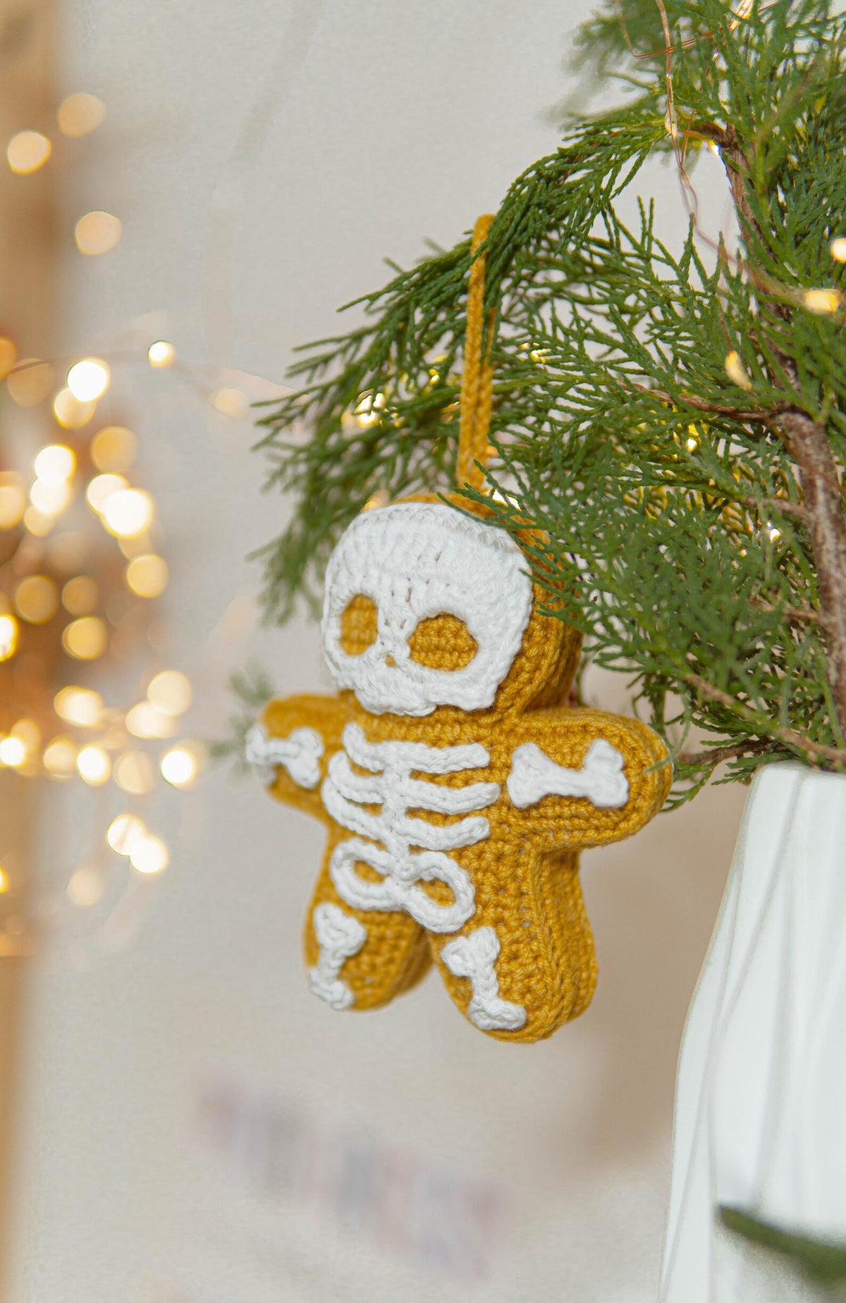 A crocheted skeleton gingerbread ornament hanging from a green evergreen tree branch with warm bokeh lights in the background.