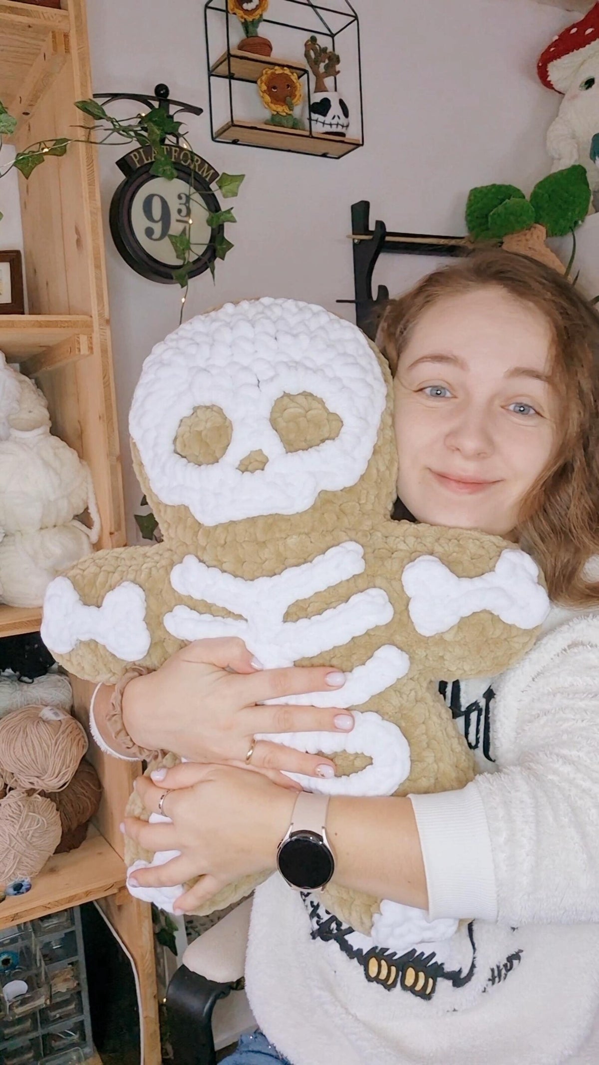 A large crocheted pillow skeleton plush toy. The background shows a cozy, cluttered craft room with shelves of yarn, knitting supplies, and other plush toys.