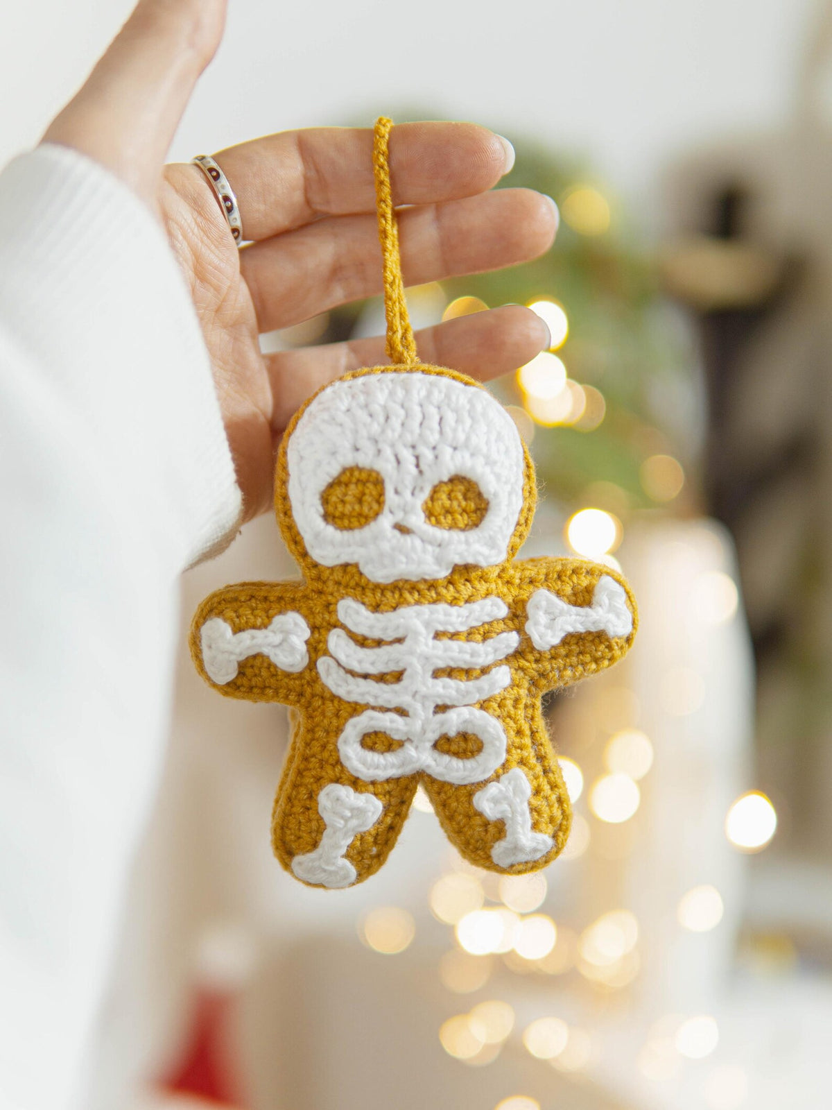 A hand holding a crocheted skeleton-shaped ornament in mustard yellow and white, with a soft blurred holiday background featuring warm bokeh lights.