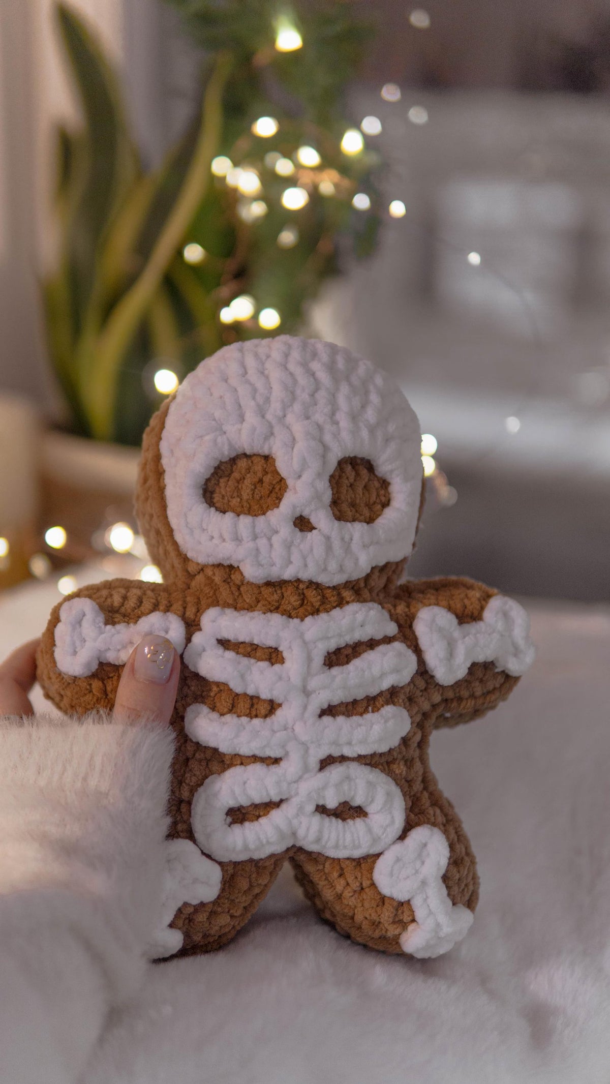 A handmade crocheted skeleton gingerbread person doll held by a hand, set against a cozy, softly blurred background with warm bokeh lights and a plant.